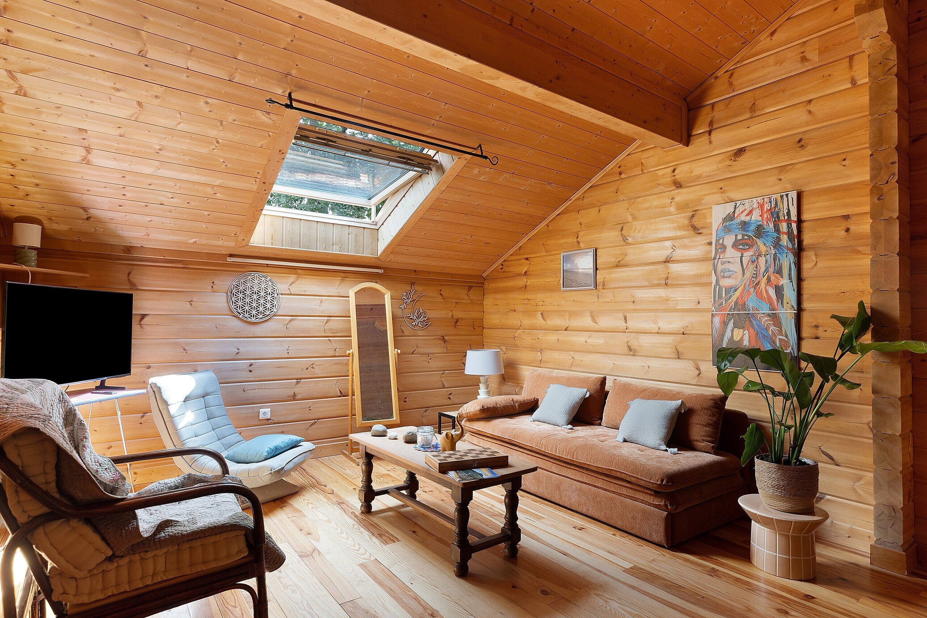 Een knus blokhutinterieur met een slaapkamer op de zolder, een houten vloer, een bank, een stoel, een tafel en een televisietoestel.