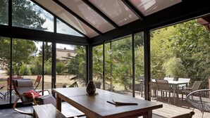 Uma estufa com mesa e bancos de madeira, amplas janelas de vidro e vista para um jardim.