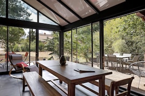 A conservatory with a wooden table and benches, large glass windows, and a view of a garden.