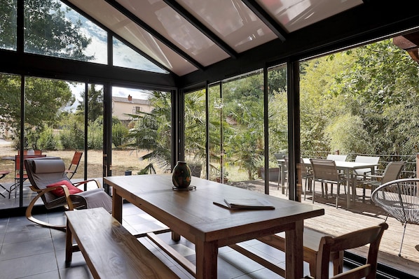 A conservatory with a wooden table and benches, large glass windows, and a view of a garden.