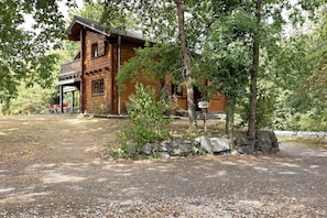 A wooden cabin with a porch, surrounded by trees and a gravel path.