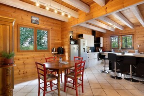 A wooden cabin interior with a dining area, kitchen, and large windows.