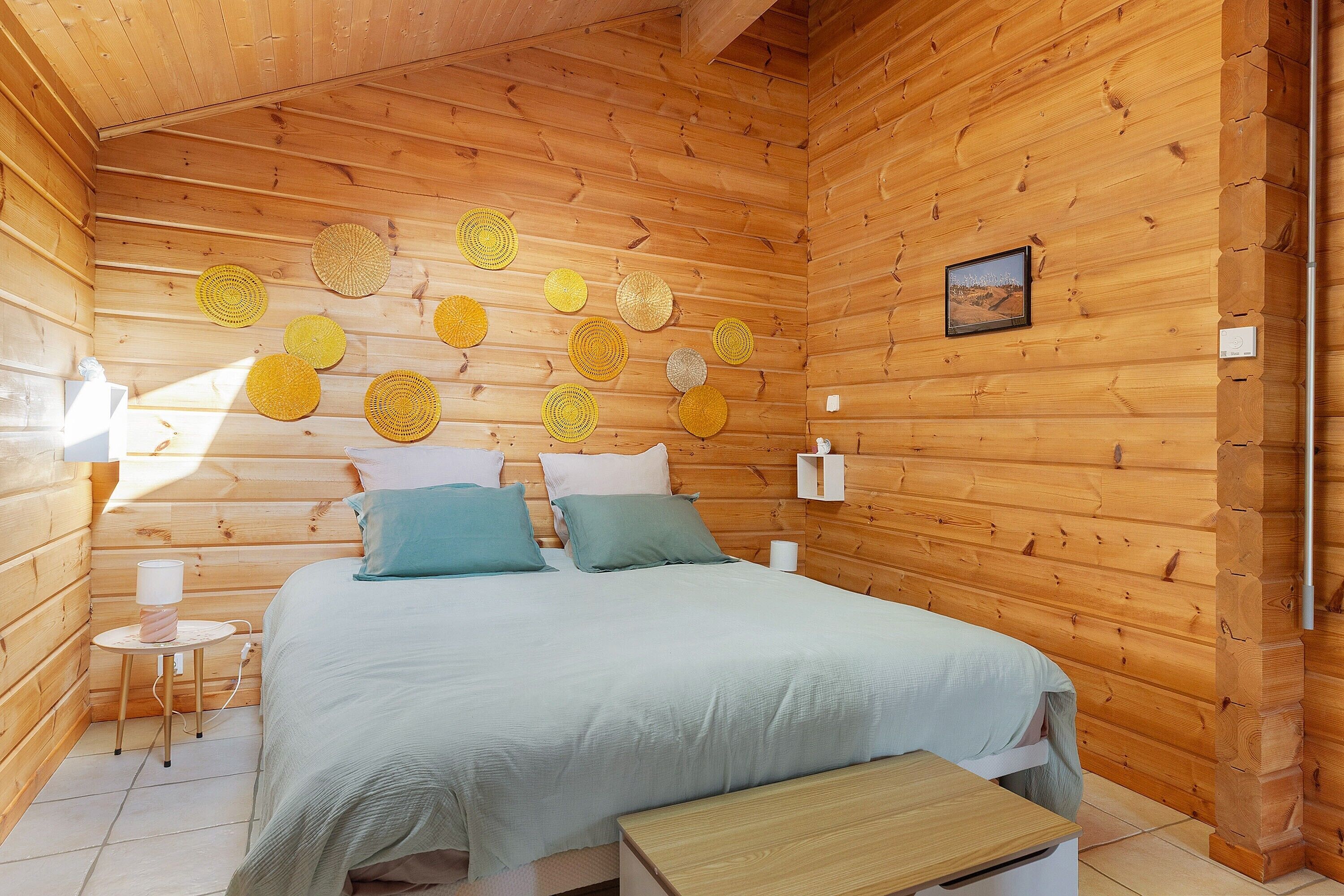 Een houten slaapkamer met een bed, kussens, een nachtkastje, een lamp en een foto aan de muur.