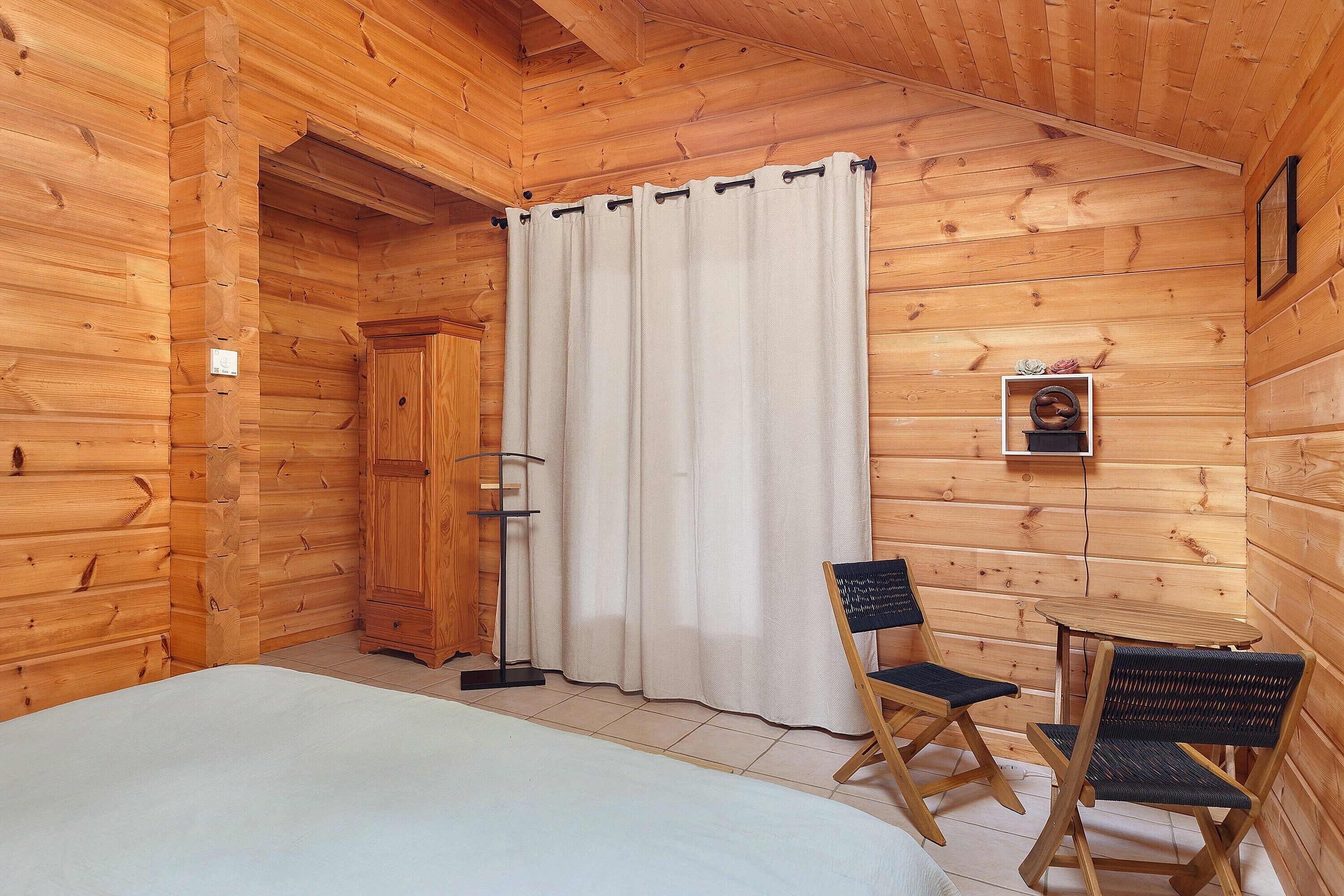 Een houten kamer met een bed, een tafeltje, twee stoelen, een gordijn en een aan de muur hangend schilderij.