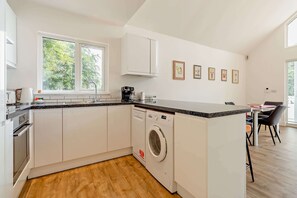 A modern kitchen with a built-in washer, a dining area with chairs, and a window with a view of trees. - Host & Stay | Robin Lodge (Burgh Castle)