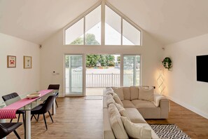 A spacious room with a high ceiling, a glass dining table, a beige sofa, and a large window. - Host & Stay | Robin Lodge (Burgh Castle)