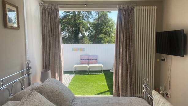 A bedroom with a bed, curtains, a TV, and a view of an outdoor area with chairs and a sign. - Moss Retreat Sleeps 2, Balcony/garden/parking/pubs (Nantwich)