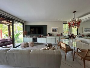 Ein modernes Wohnzimmer mit einer Couch, einem Esstisch und Stühlen, einer Küchenzeile mit Fernseher und großen Fenstern mit Blick auf Grünflächen.