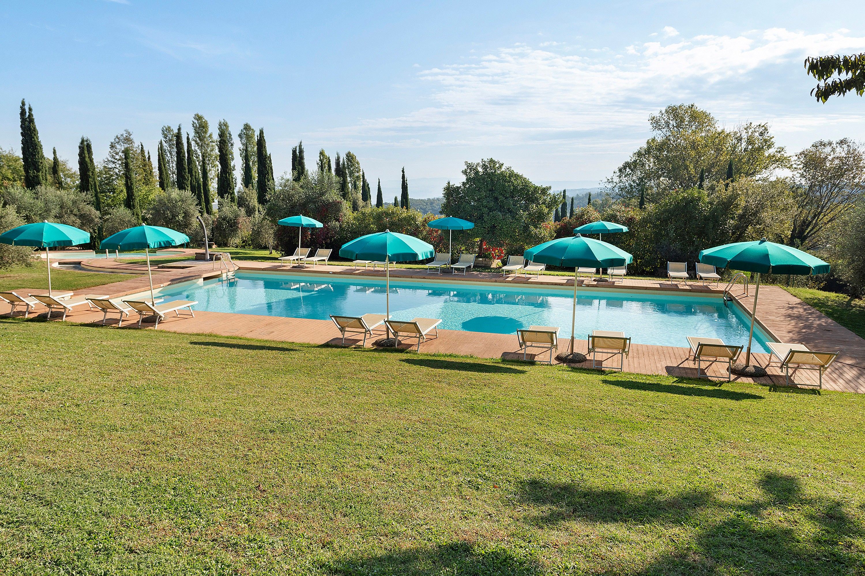 Une piscine entourée de pelouse et d’arbres, avec des chaises longues et des parasols.
