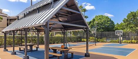 An outdoor basketball court with a covered pavilion, picnic tables, and a basketball hoop.