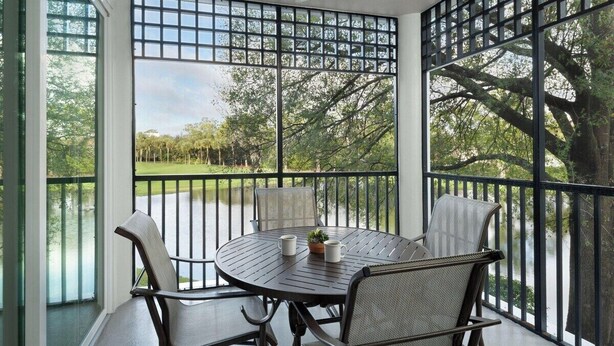 Un porche techado con una mesa redonda y cuatro sillas, una pequeña planta en maceta y una vista de un paisaje verde a través de una celosía metálica.