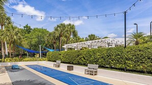 Un espacio exterior soleado con una cancha de shuffleboard, asientos y un edificio al fondo.