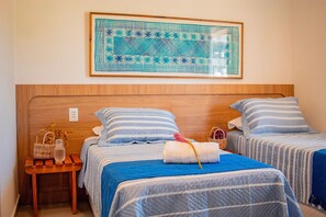 Ein Hotelzimmer mit zwei Betten, einem Holzkopfstück, einem an der Wand hängenden Kunstwerk und einem kleinen Holztisch mit Korb und Vase.