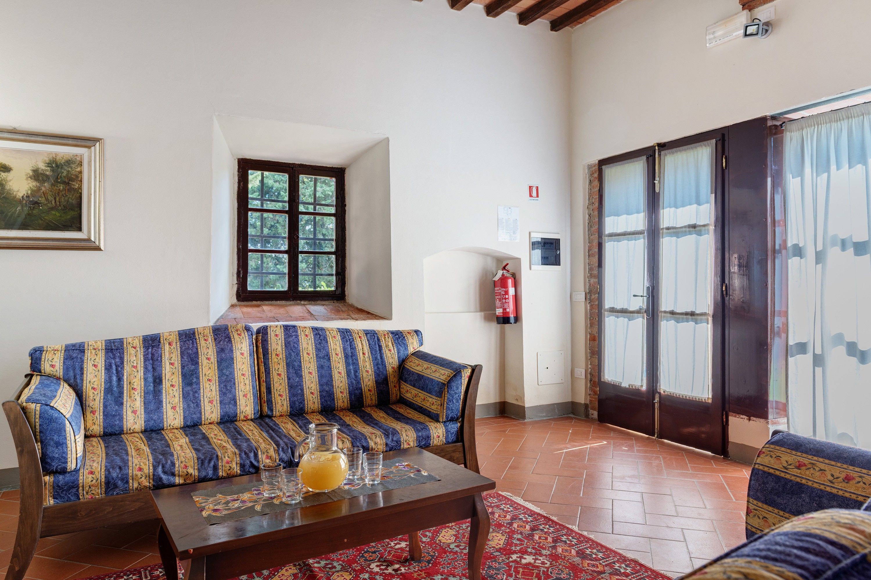 A living room with a sofa, a coffee table with a pitcher and glasses, a fire extinguisher, and a door with curtains.
