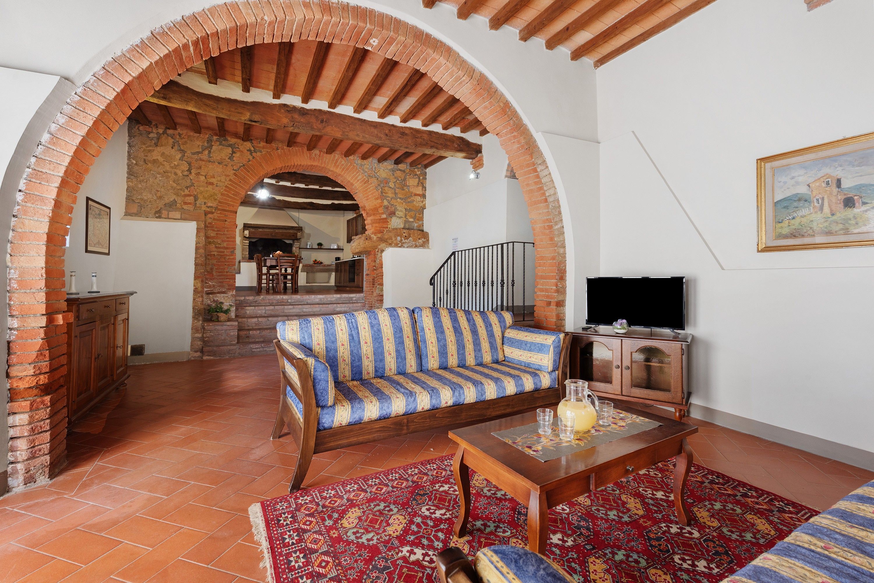 A living room with a sofa, a coffee table, and a TV. There is a fireplace, a staircase, and a kitchen in the background.