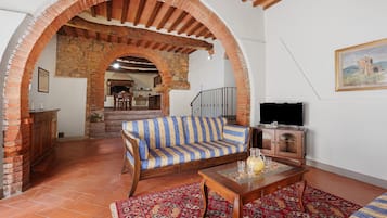 A living room with a sofa, a coffee table, and a TV. There is a fireplace, a staircase, and a kitchen in the background.