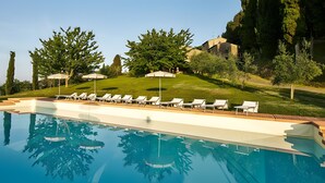 Una piscina con lettini e ombrelloni, circondata da alberi e una collina.
