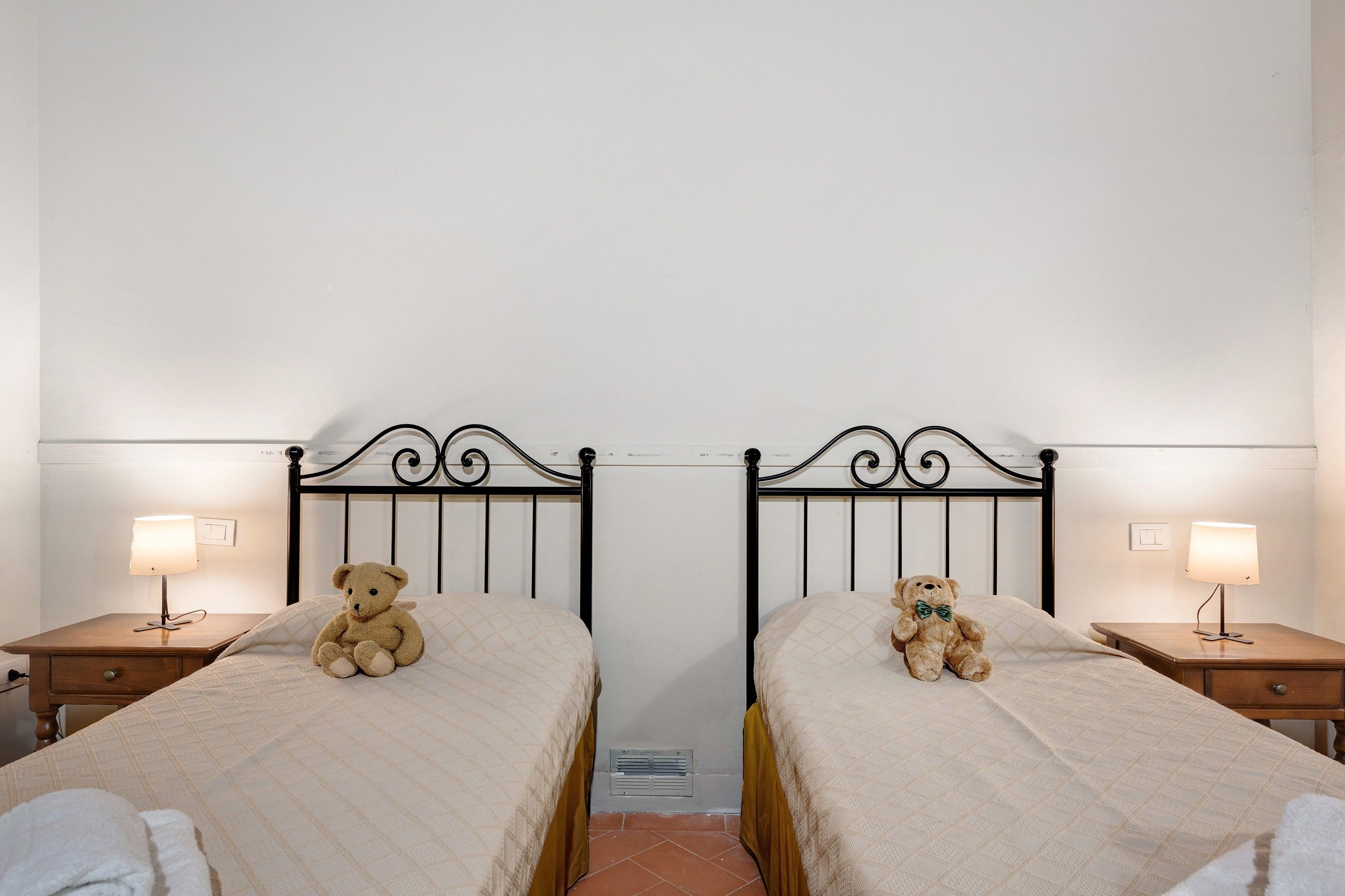 A room with two beds, each with a headboard, a bedside lamp, and a teddy bear on the bed.