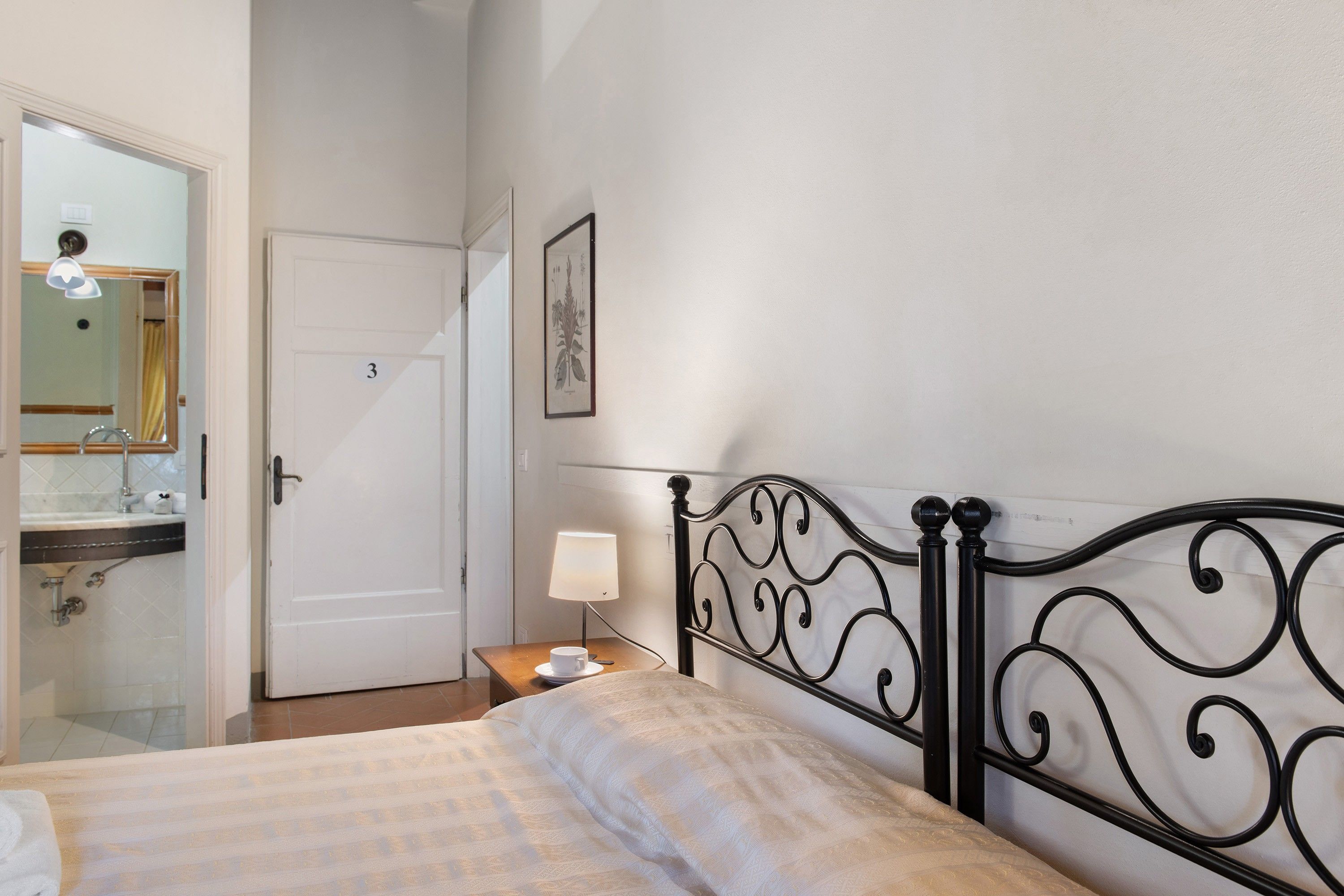A bedroom with a bed, a bedside table with a lamp, a bathroom with a mirror, and a door.