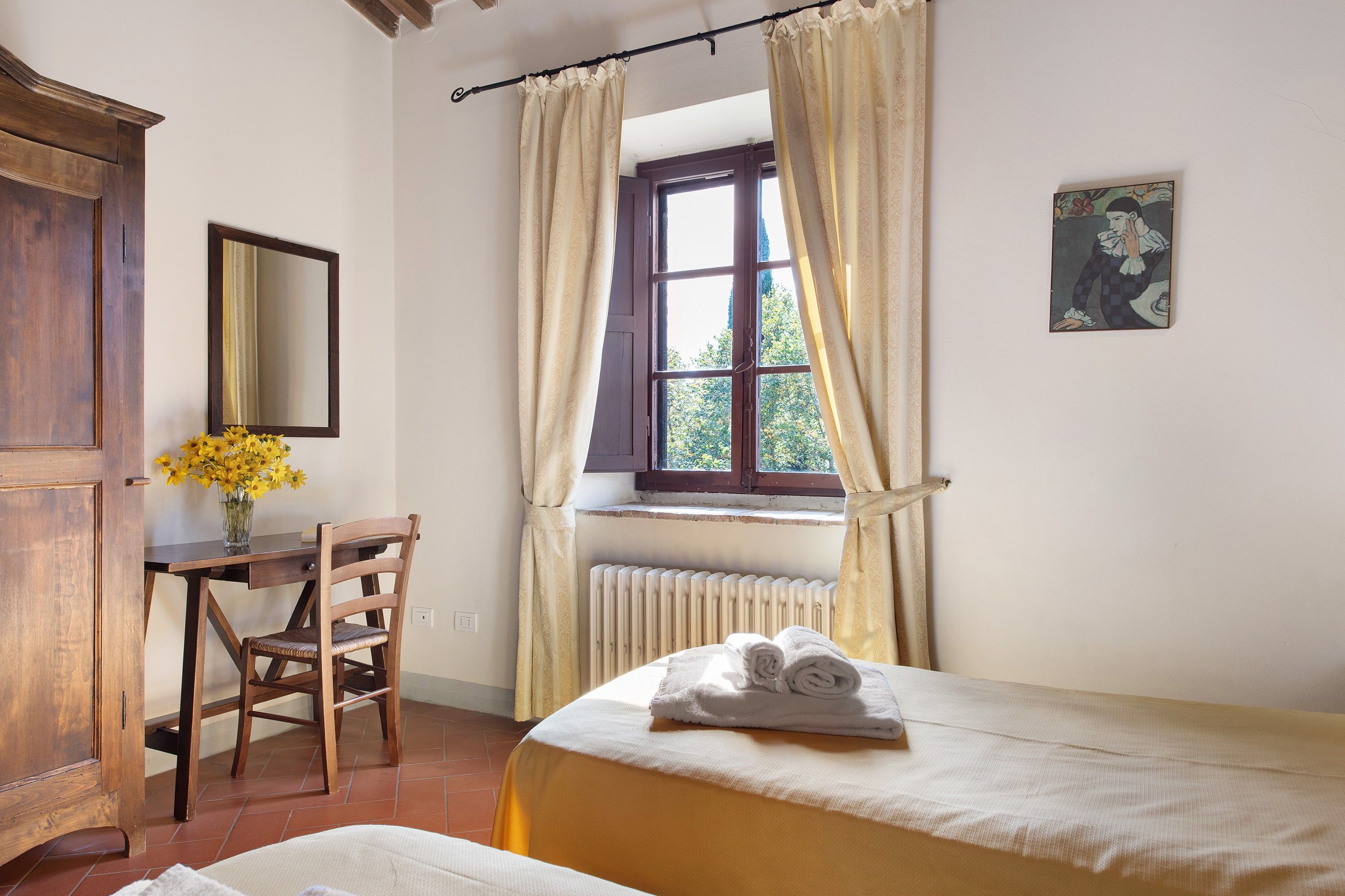 A bedroom with a bed, a chair, a desk, a window with curtains, a radiator, and a painting on the wall.
