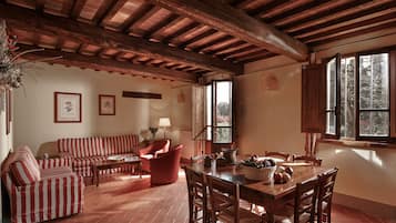 Ein rustikales Interieur mit Holzbalkendecke, einem Esstisch mit Stühlen, einer Couch und einem roten Sessel.