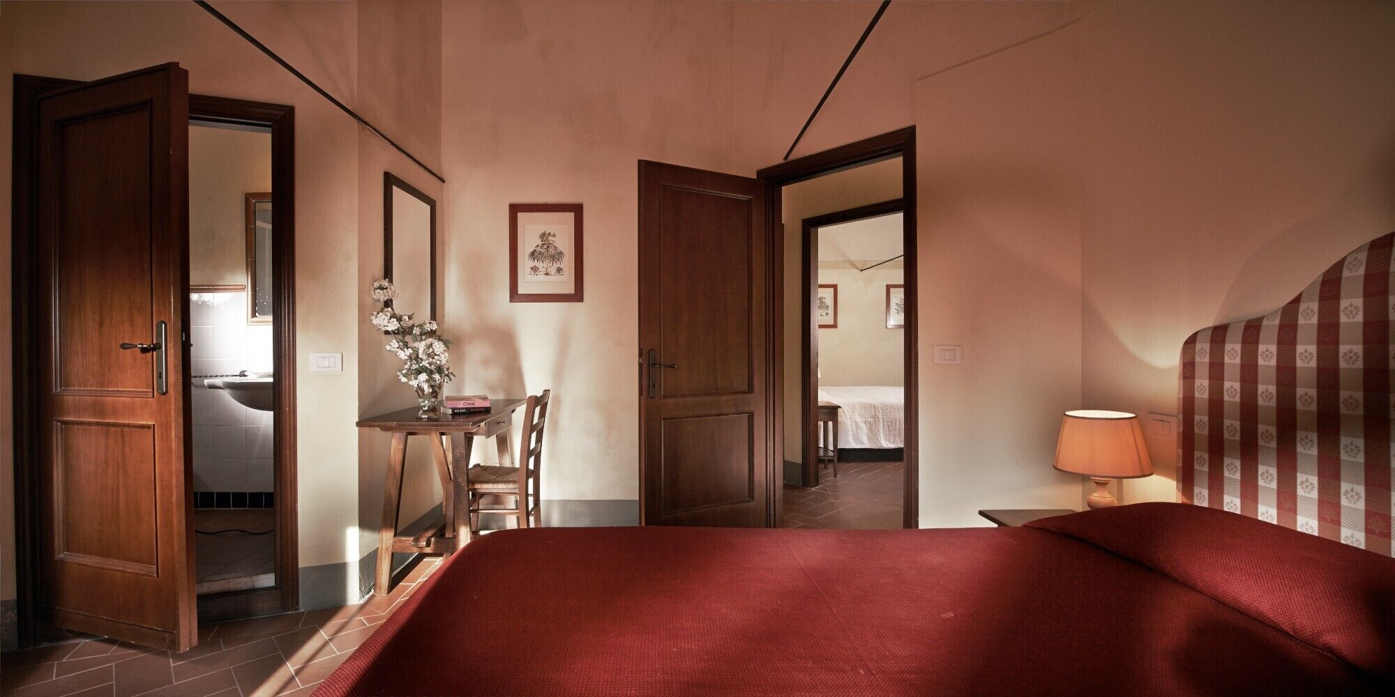 Une chambre d’hôtel avec un lit, un bureau et une chaise, une lampe, et une salle de bain visible à travers une porte ouverte.