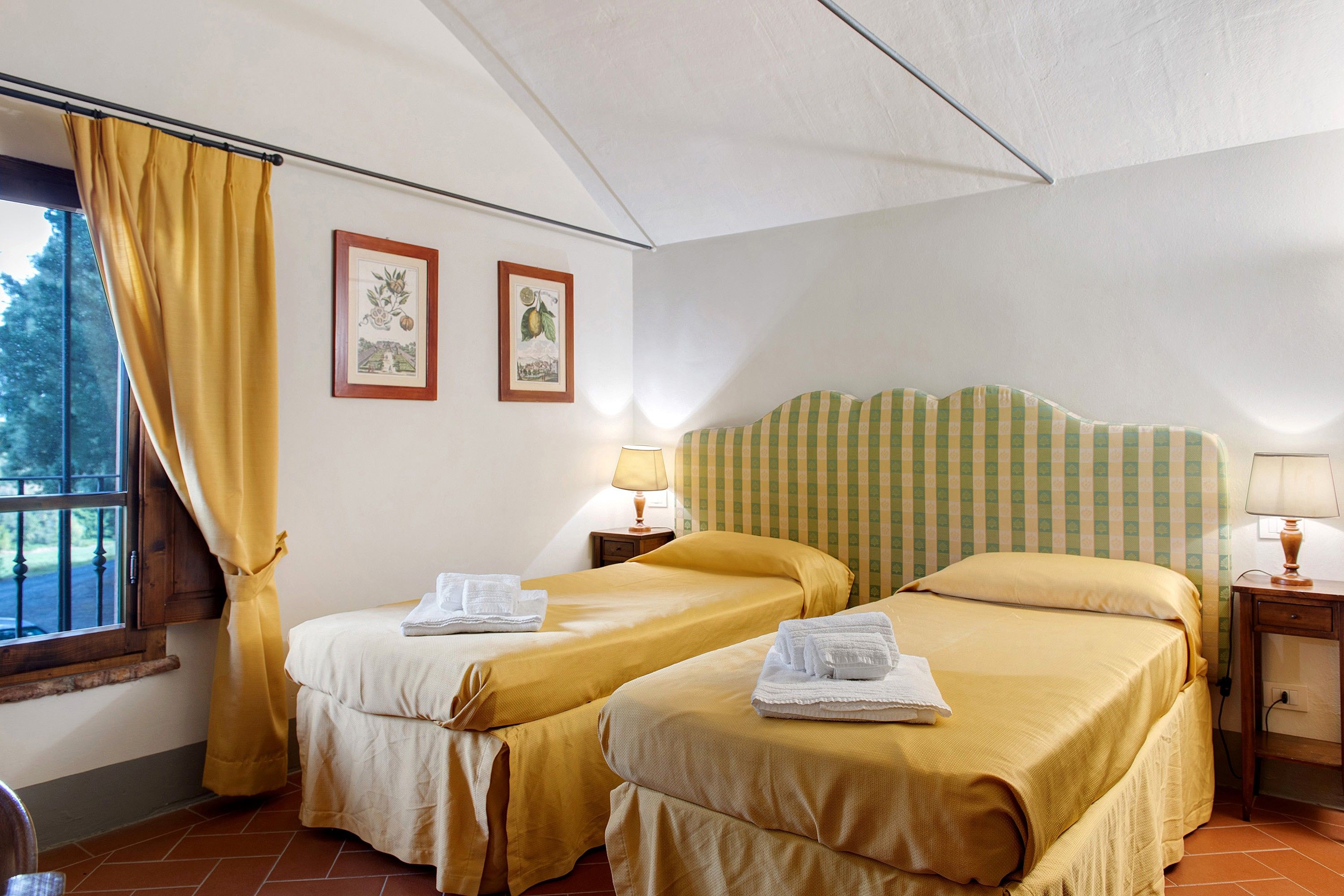 Habitación de hotel con dos camas, una ventana con cortinas, una lámpara y dos cuadros en la pared.