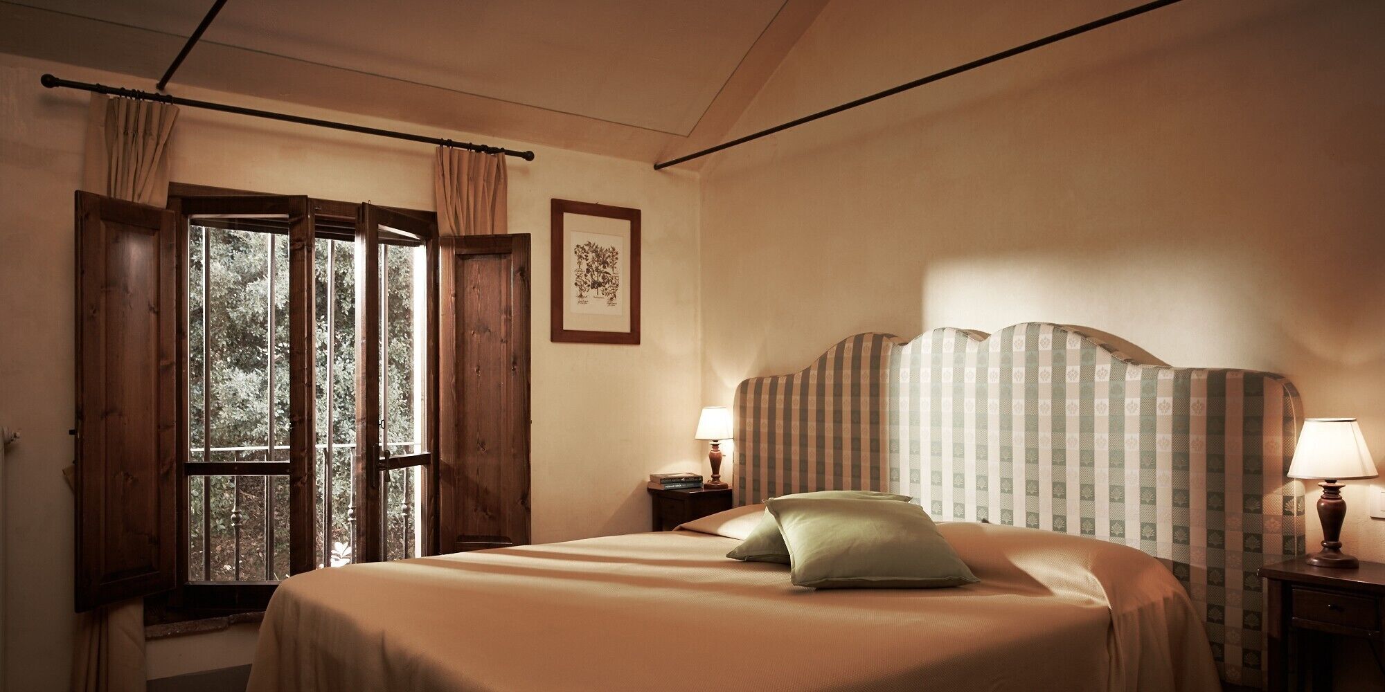 Une chambre d’hôtel avec un lit, une table de chevet, une lampe, une fenêtre avec des rideaux et un tableau encadré au mur.