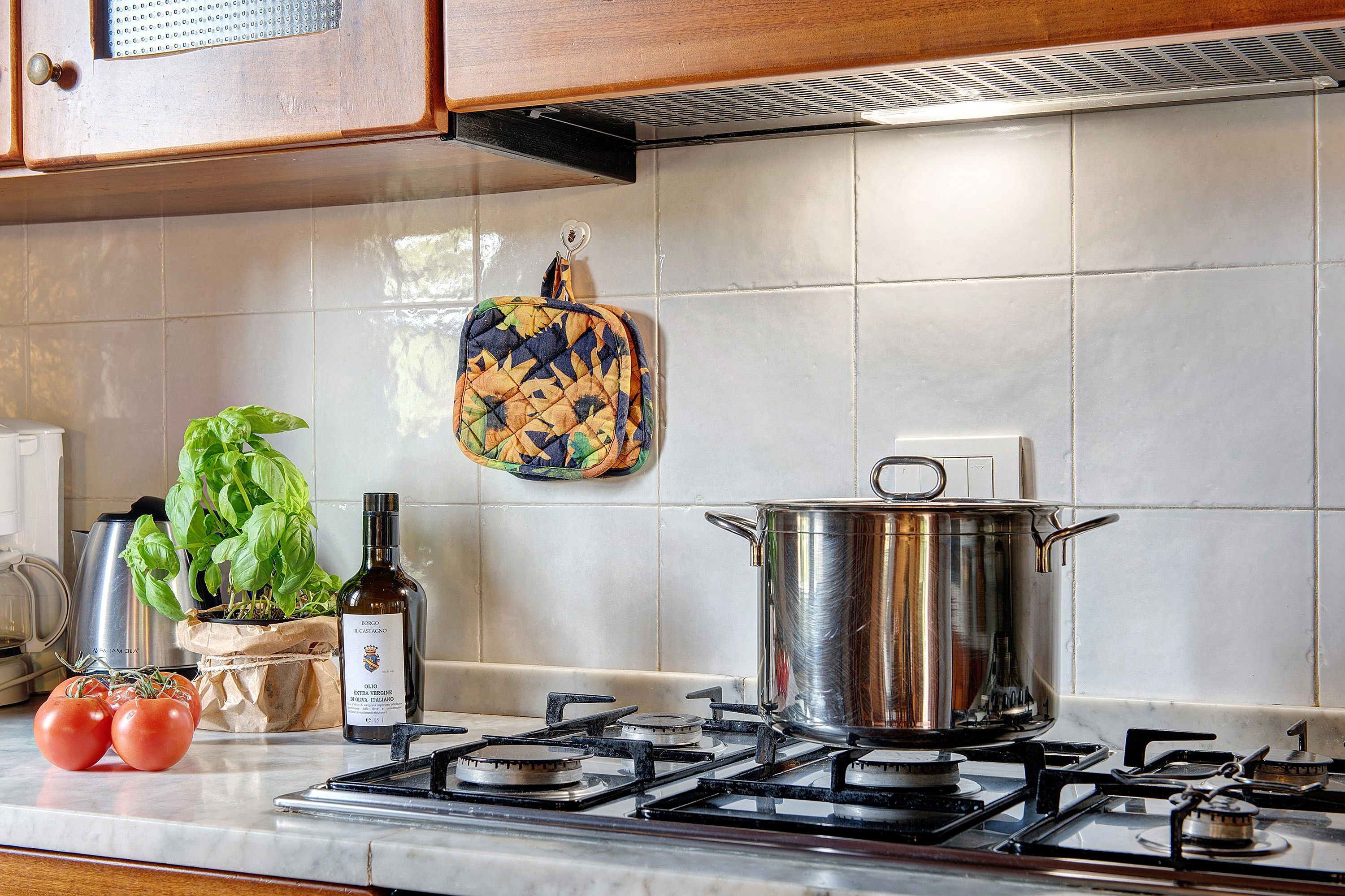 Una cocina con estufa a gas, una olla, una botella de aceite de oliva, tomates, albahaca y un salvador para ollas colgado.