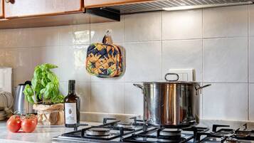Una cocina con estufa a gas, una olla, una botella de aceite de oliva, tomates, albahaca y un salvador para ollas colgado.