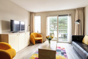 A modern living room with a flat-screen TV, a yellow armchair, a black sofa, and a coffee table with a vase of flowers. - Comfortable vacation apartment on Ameland with access to a pool; dogs welcome (Hollum)