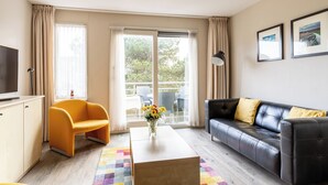 A modern living room with a sofa, armchair, TV, and a view through the glass door. - Comfortable vacation apartment on Ameland with access to a pool; dogs welcome (Hollum)