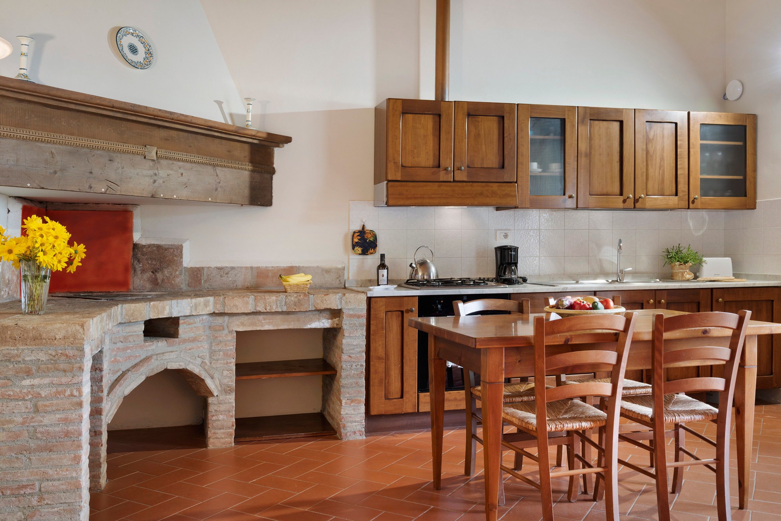 Een keuken met een bakstenen open haard, houten kastjes en een eethoek met stoelen.