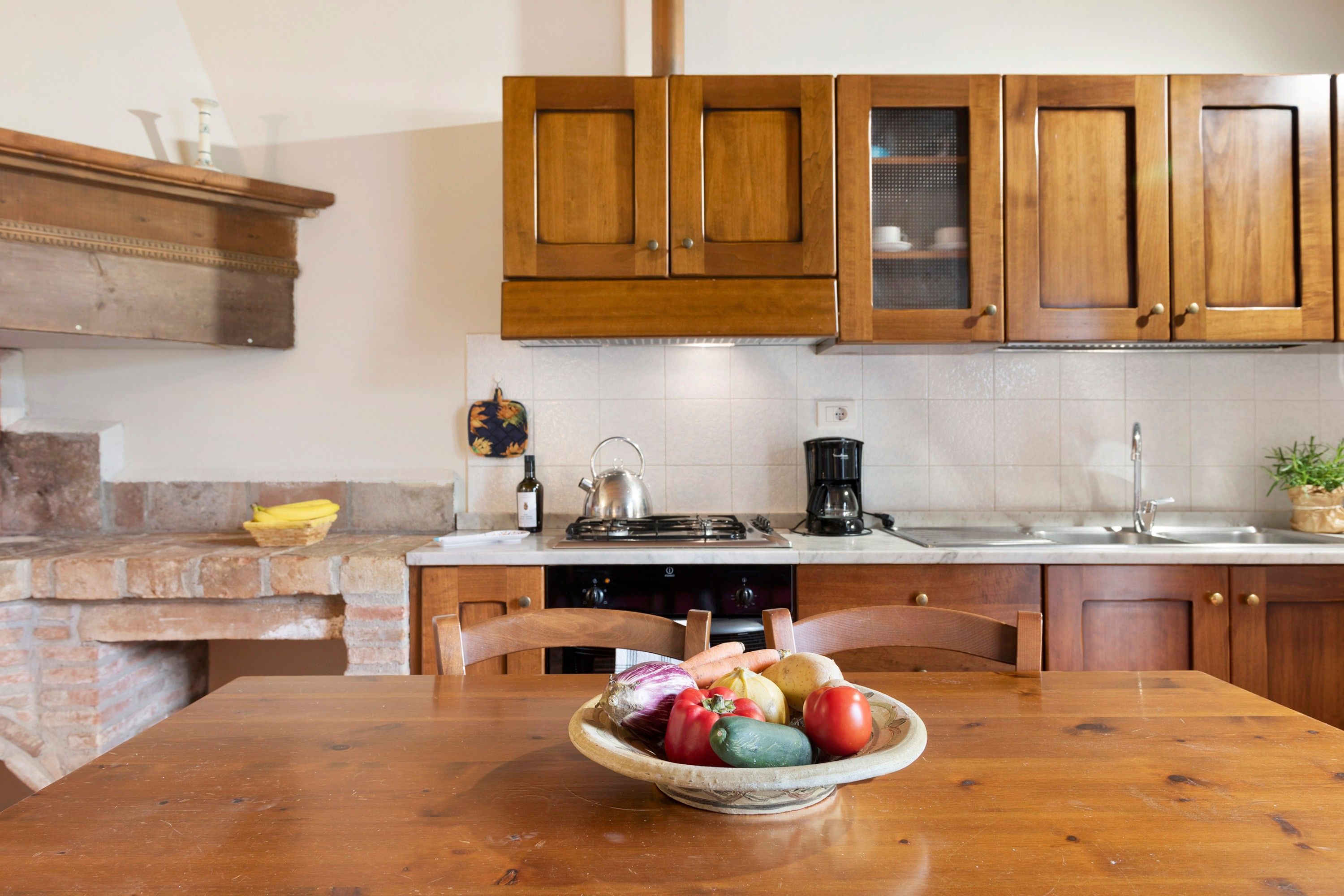 Una cocina con gabinetes de madera, salpicadero de ladrillo, una estufa a gas y un tazón de verduras sobre la mesa.