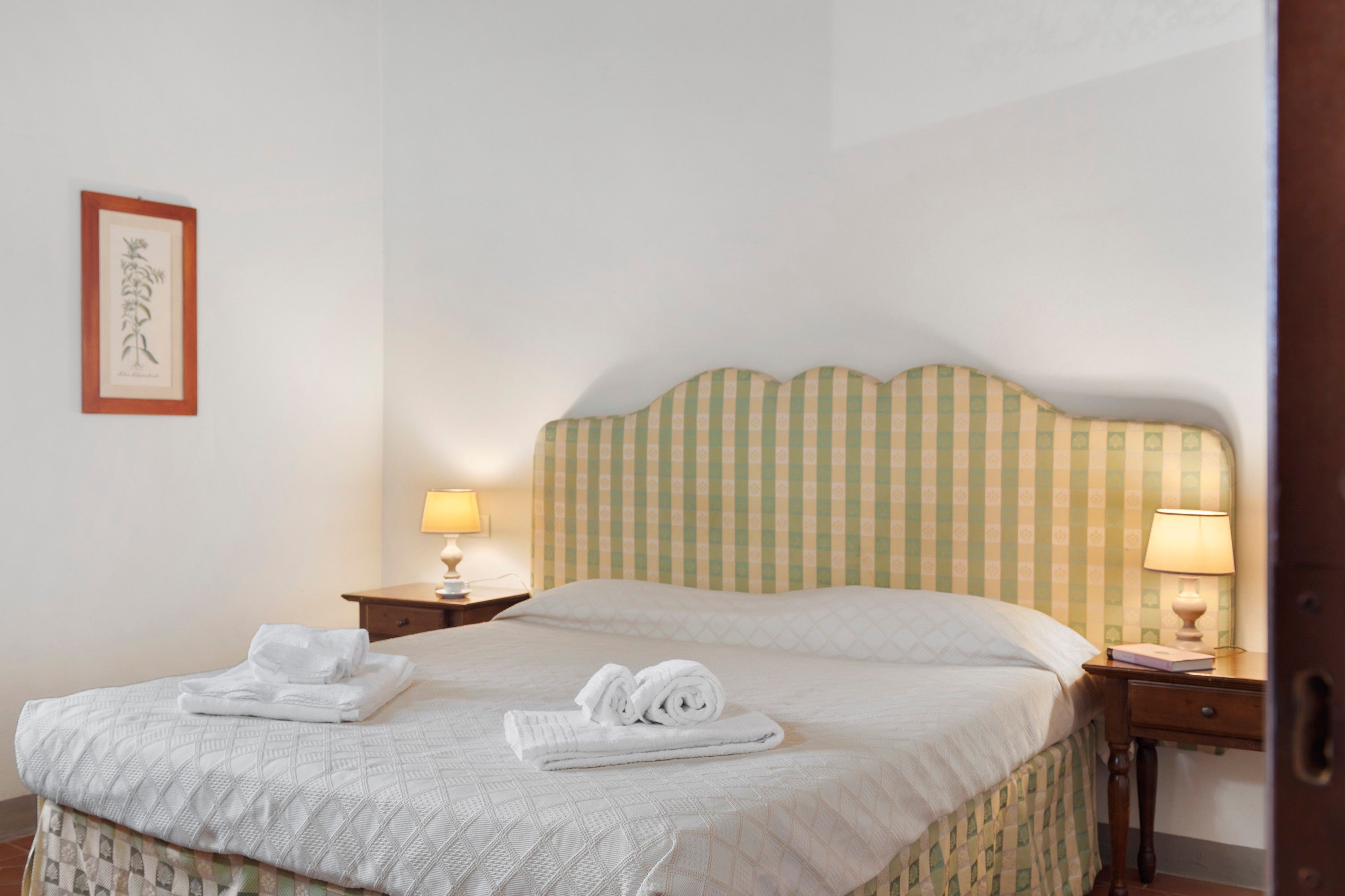 Een hotelkamer met een bed, twee nachtkastjes, een lamp en een ingelijste botanische afbeelding aan de muur.