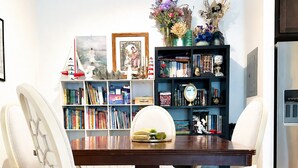 Un coin repas avec une table en bois, des chaises à revêtement blanc et une bibliothèque remplie de livres et d’objets décoratifs.