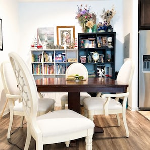 A dining area with a wooden table, white upholstered chairs, and a bookshelf filled with books and decorative items. - Summer Heaven Retreat near Universal Studious, Disney (Lake Buena Vista)