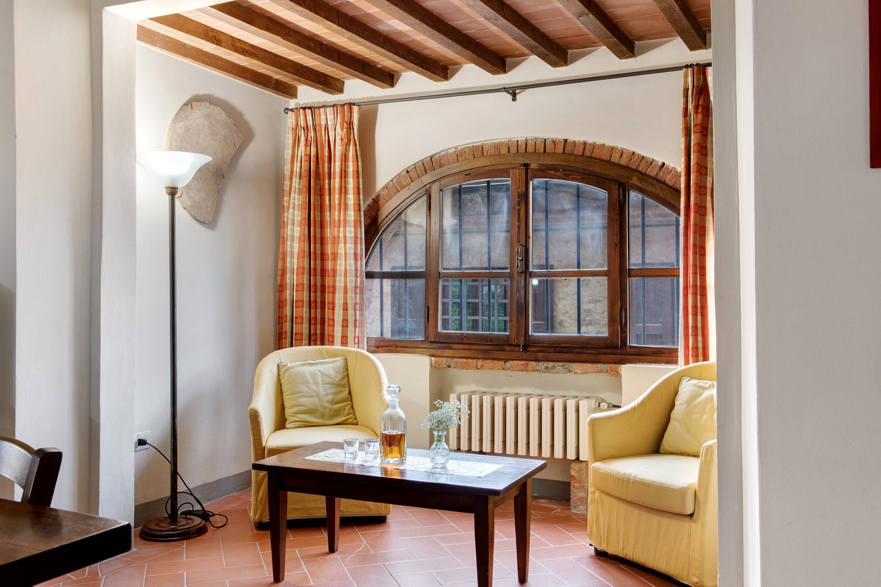 A room with a large arched window, two armchairs, a table with a bottle and glasses, a floor lamp, and a radiator.