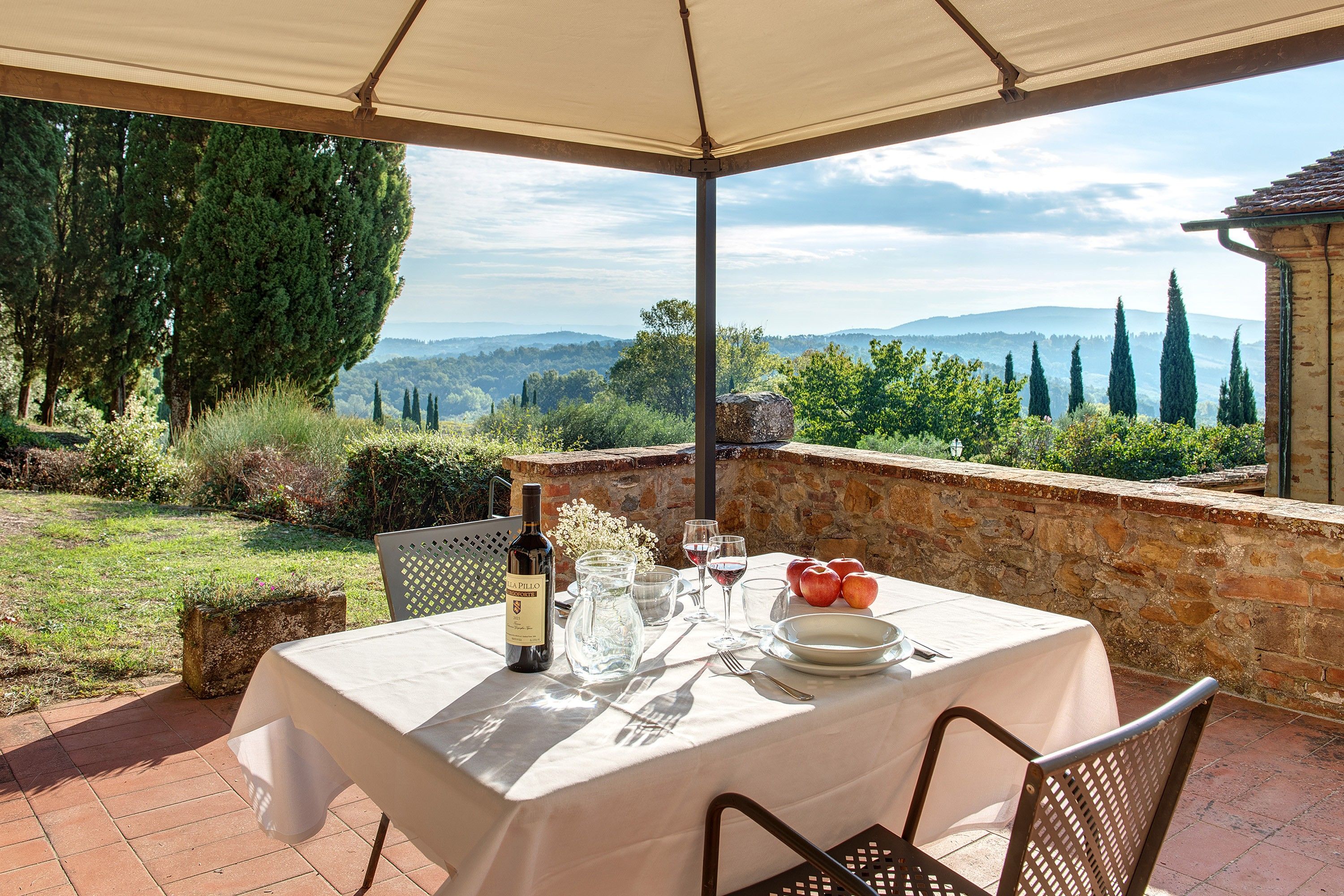 Un área de comedor al aire libre con una mesa preparada para dos personas, una botella de vino y una jarrón. El entorno incluye un muro de piedra y vistas al campo.