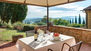 Un área de comedor al aire libre con una mesa preparada para dos personas, una botella de vino y una jarrón. El entorno incluye un muro de piedra y vistas al campo.