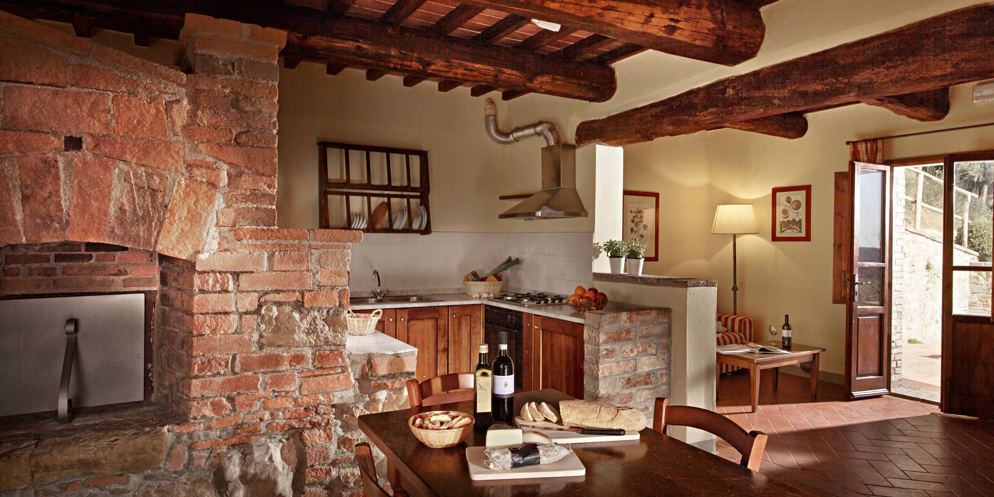 Une cuisine rustique aux murs en briques apparentes, avec des poutres en bois, une cheminée en pierre et un coin repas où une table est dressée pour le repas.
