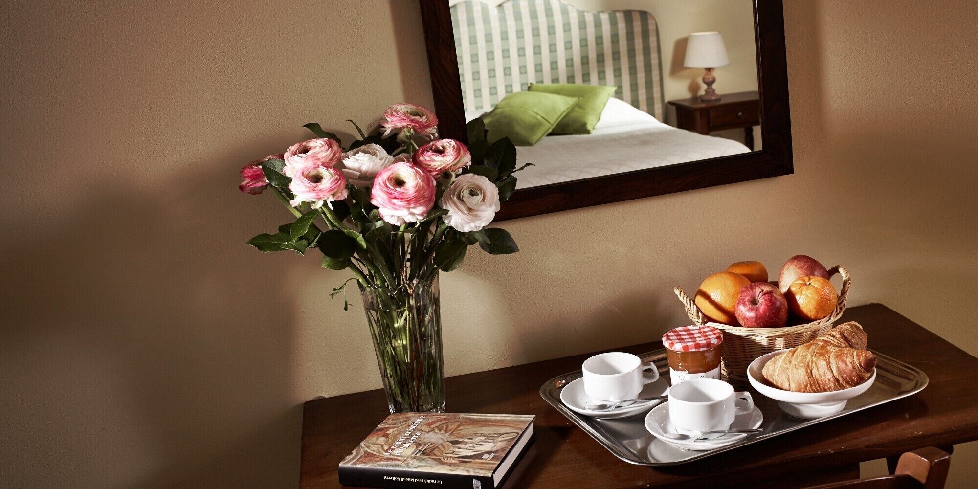 Ein Hotelzimmer mit einem Bett, einem Nachttisch mit Blumen, einer Ablage mit Croissants, Äpfeln, Orangen und einem Buch.