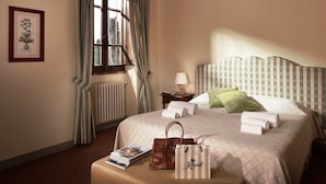 Un lit bien fait, avec des oreillers, une table de chevet avec une lampe, une illustration botanique encadrée accrochée au mur et une fenêtre avec des rideaux.
