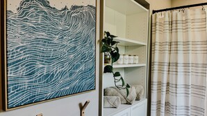 A bathroom with a wooden wall-mounted towel rack, a framed artwork depicting a mountain landscape, and a shelf with cotton balls and a plant.