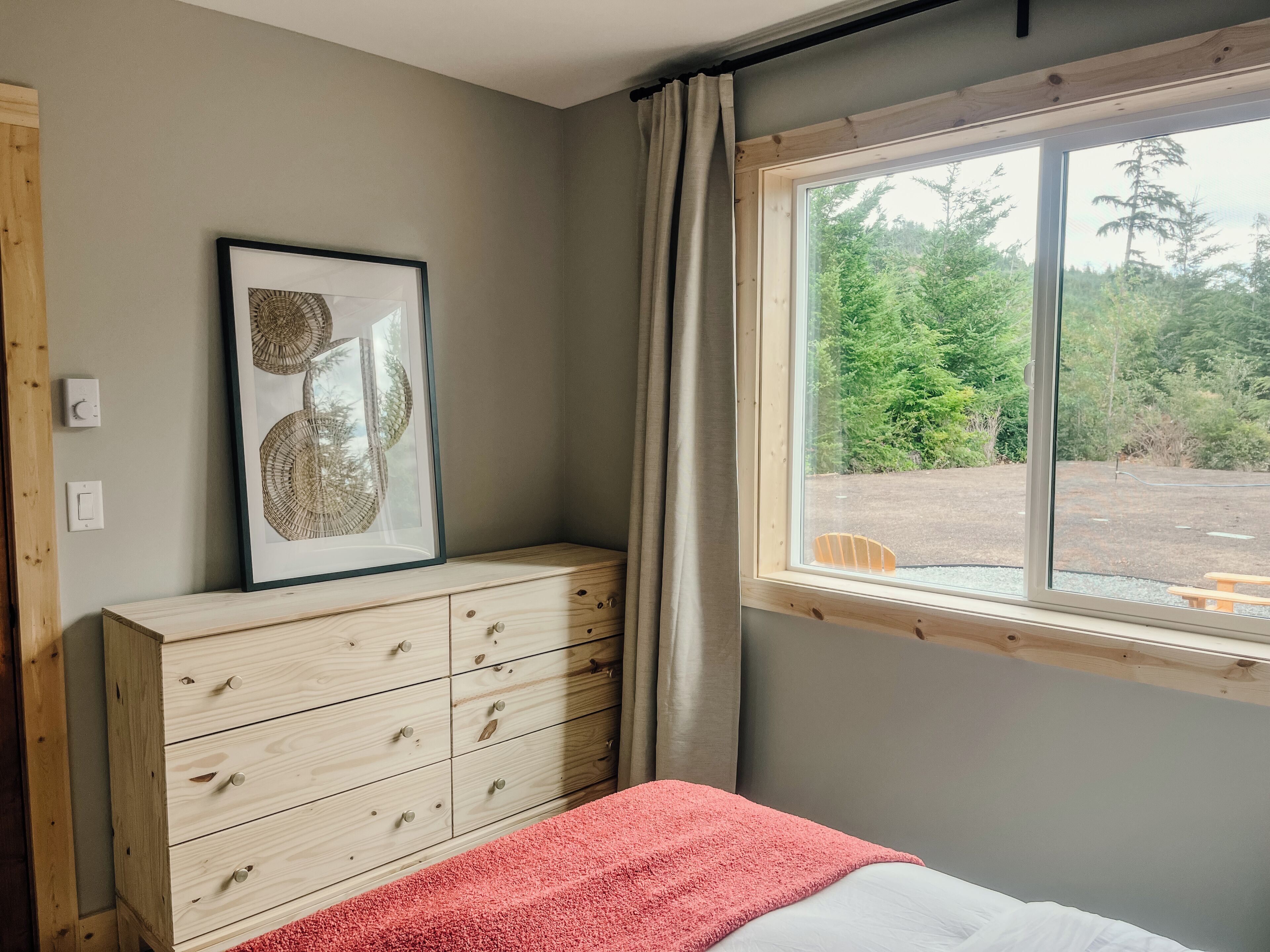 Une chambre à coucher avec un lit, une commode, un tableau encadré, une fenêtre avec des rideaux et une vue sur la verdure à l’extérieur.