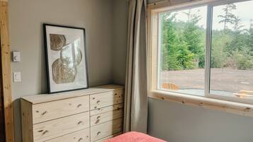 Une chambre à coucher avec un lit, une commode, un tableau encadré, une fenêtre avec des rideaux et une vue sur la verdure à l’extérieur.