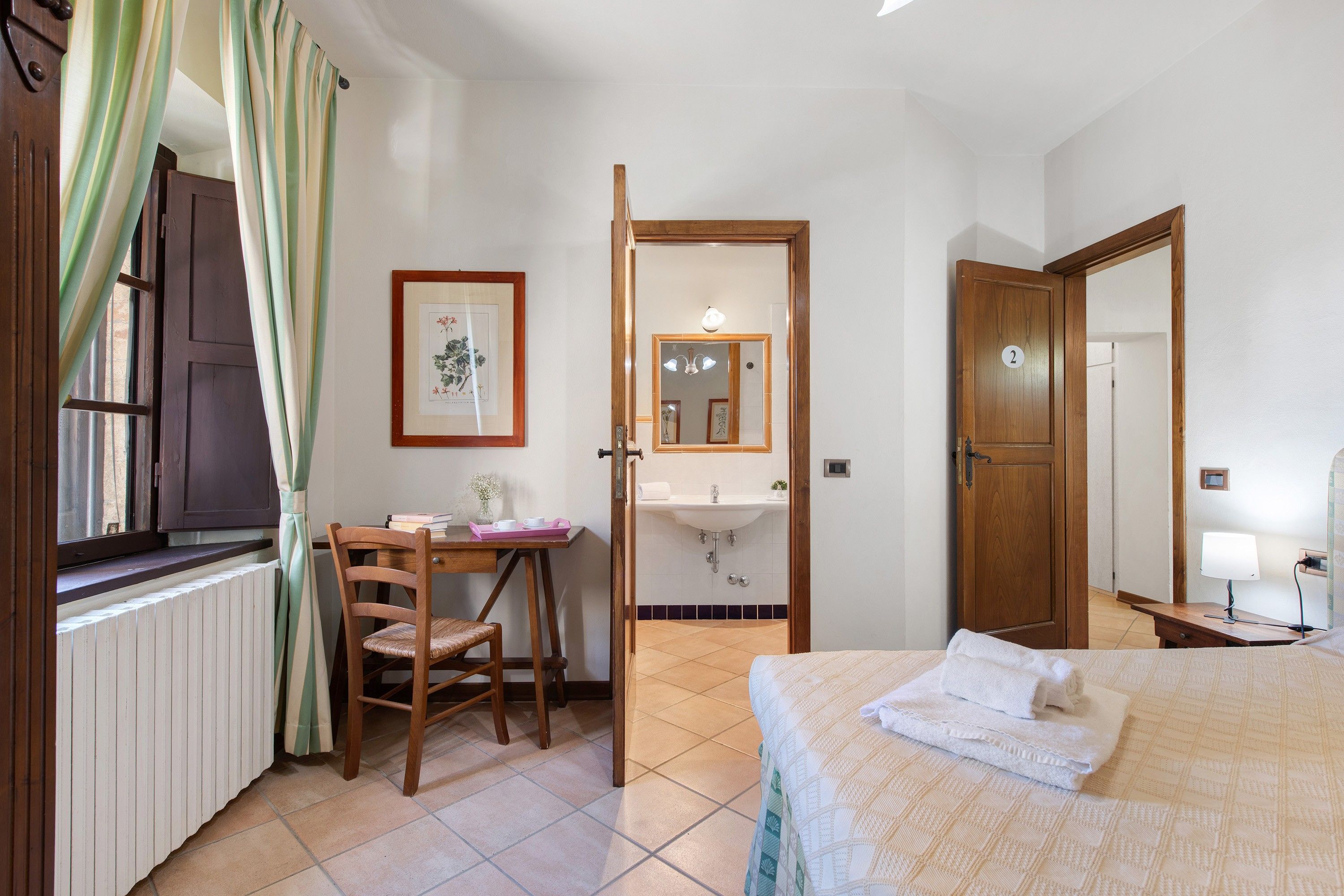 Un dormitorio con cama, un escritorio con silla, una ventana con cortinas, un baño con lavamanos y espejo, y una puerta con un número.