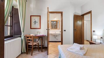 Un dormitorio con cama, un escritorio con silla, una ventana con cortinas, un baño con lavamanos y espejo, y una puerta con un número.