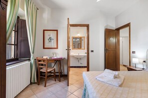 Un dormitorio con cama, un escritorio con silla, una ventana con cortinas, un baño con lavamanos y espejo, y una puerta con un número.