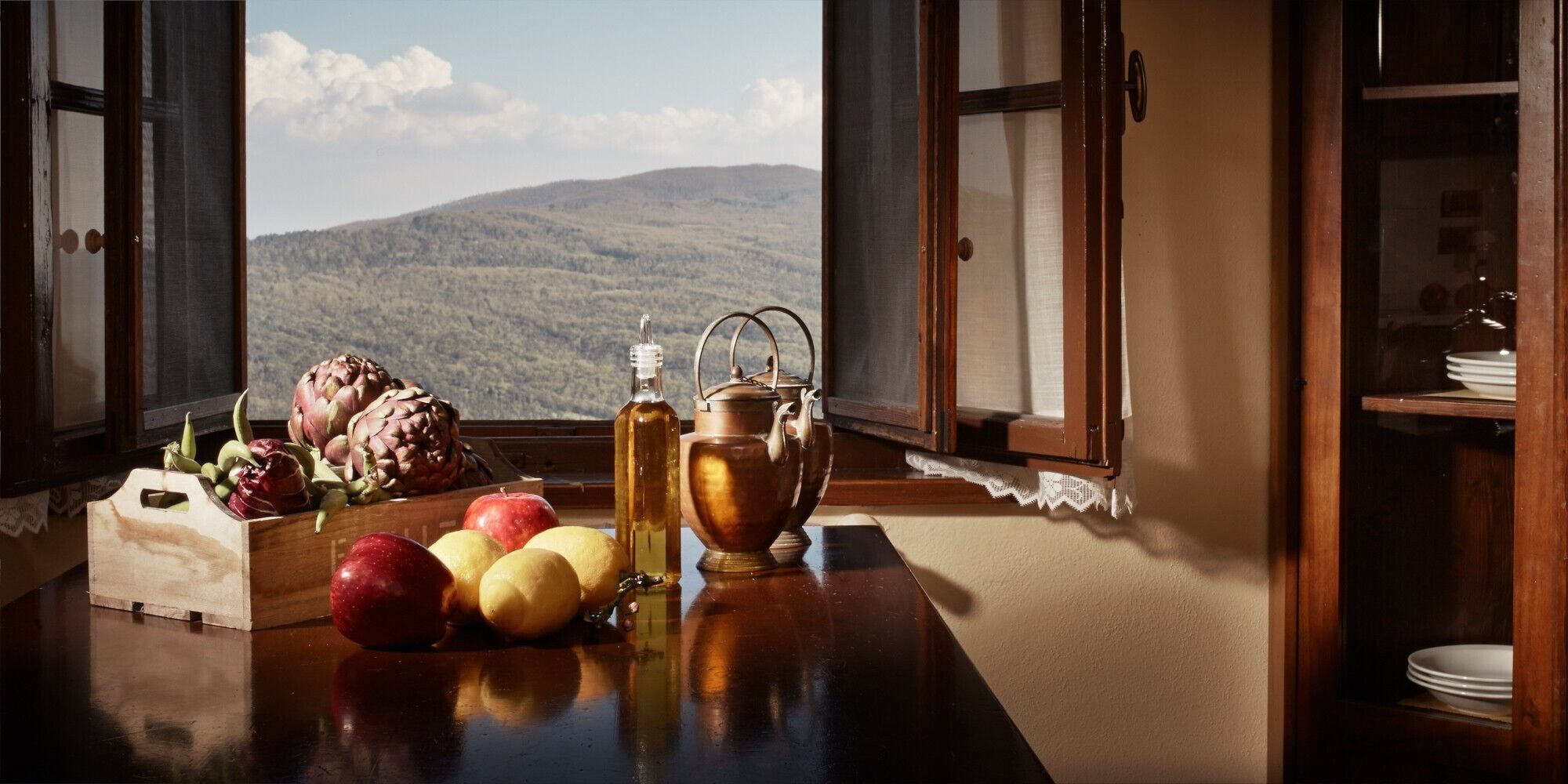 Ein Tisch mit Früchten, einer Flasche und einem Teekessel vor einem Fenster mit Blick auf die Berge.